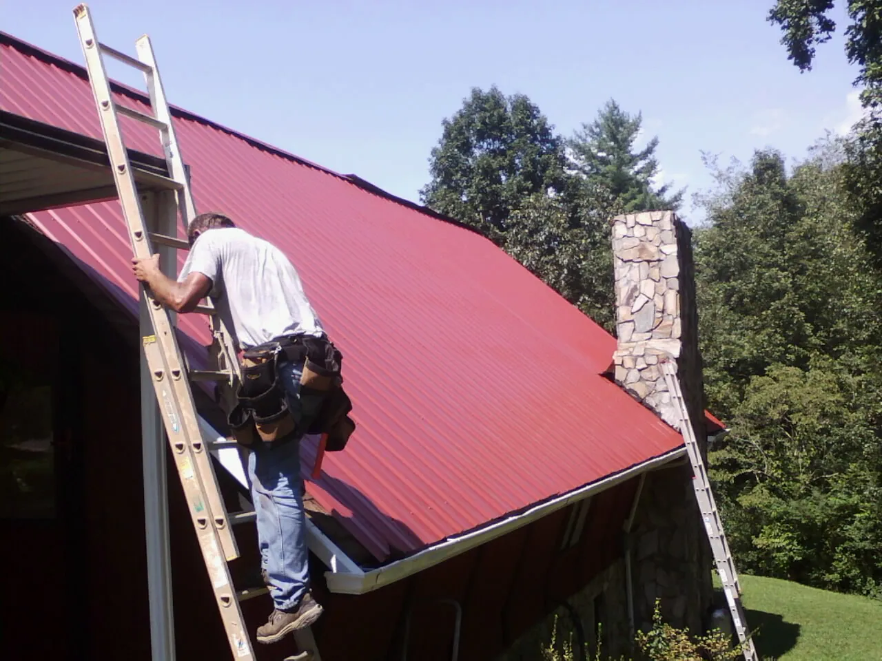 Roofer on a ladder beside a red metal roof and stone chimney — Soffit & Fascia Repair by Roofing Brothers in Gold Bar